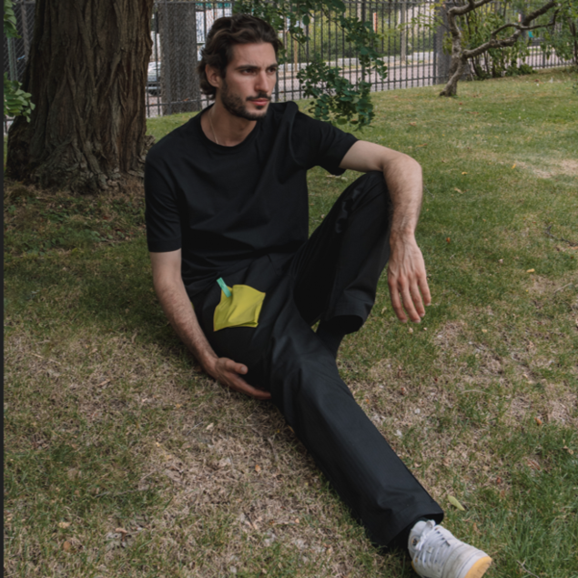 A male model seated on grass, leaning against a tree, dressed in all black with a distinctive yellow pocket on his pants.
