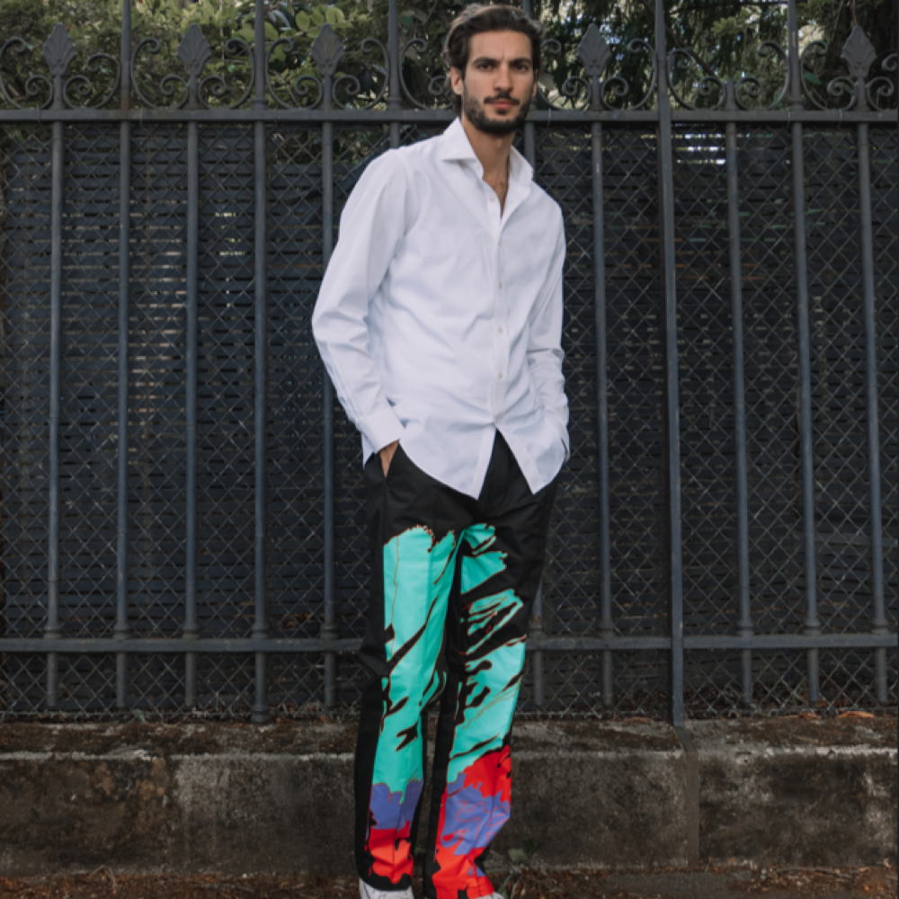 Person standing by a fence, wearing a white shirt and colorful patterned trousers.