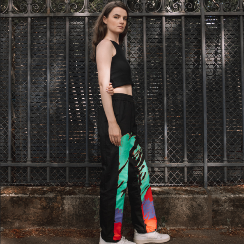 Individual leaning against a fence, wearing a black crop top and pants with a colorful pattern on one leg.
